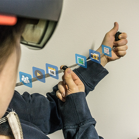 The picture shows the ARCord prototype in action. A woman is wearing a hoodie and a HoloLens. She pulls the hoodie cord in front of her and sees holographic overlays that she controls by moving the hood cord lock.