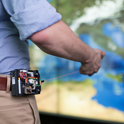 The image shows how a person wears the belt-worn elastic controller in front of a large display wall showing a map visualization. The user pulls the string towards the display.