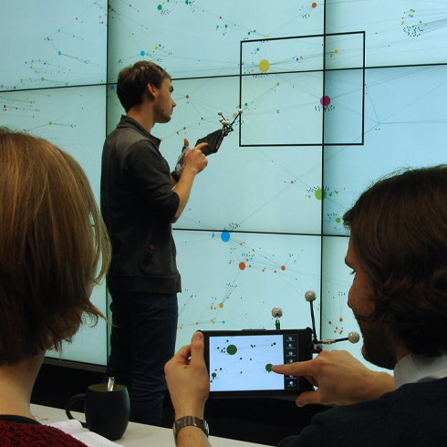 The photo shows people interacting with a large-scale display wall and mobile devices to explore a graphical dataset. In detail, a man touches the wall while a woman and another man sit on a table and use the tablets as pointing devices.